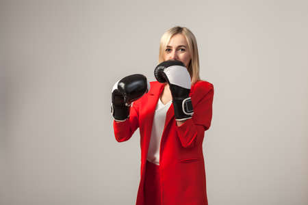 Young beautiful white blond girl in a bright red strict suit with a jacket and white blouse standing in a pose with boxing gloves on a white isolated backgroundの写真素材