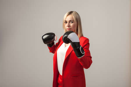 Young beautiful white blond girl in a bright red strict suit with a jacket and white blouse standing in a pose with boxing gloves on a white isolated backgroundの写真素材