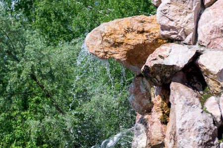 A small waterfall sprinkles drops of water down from a stone in the form of a rock against the background of green trees. Decorative designer fountain.の写真素材