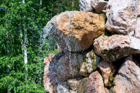 A small waterfall sprinkles drops of water down from a large stone in the form of a rock against the background of green trees. Decorative designer fountain.の写真素材