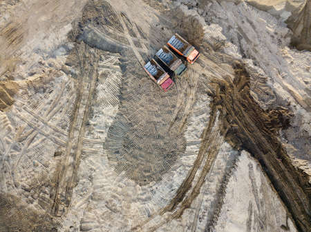 Aerial top view of three tip trucks standing next to each other awaiting loading of sand and soil on a construction site with a sand surface and a country road to the destination among uneven groundの写真素材