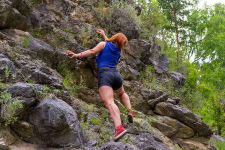 Young red-haired women climber in sports clothes climbs on rocks to the top in mountains of Altai...