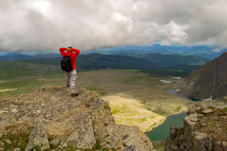 Guy in a red sweatshirt with a backpack is standing with his hands behind head on the edge of the cliff,in front of him is a colorful view of mountains,lakes with glaciers.Traveler completes the routeのeditorial素材