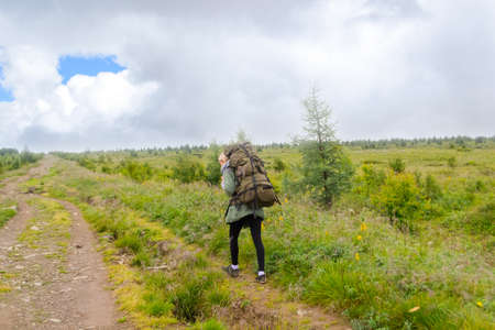 Young woman travel with big green backpack and fishing rod in it go on the road, along the green...