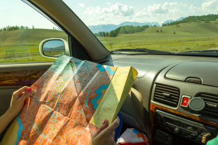 The girl rides in the car in the passenger seat and looks at the paper map looking for the desired...