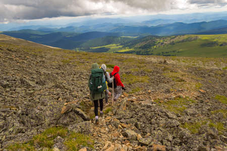 Three tourists two girls and one boy with backpacks go down the hill on the rocks towards the green fields, adventures under the blue sky and clouds in altai mountains named sarlykの写真素材