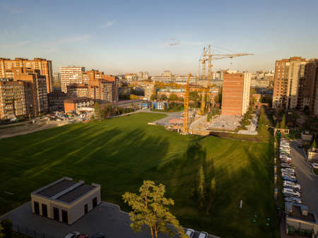 Aerial view of landscape with an elite building in the center of the city with skyscraper and three large yellow cranes with a large lawn of green grass in the fall during sunset on a clear dayのeditorial素材
