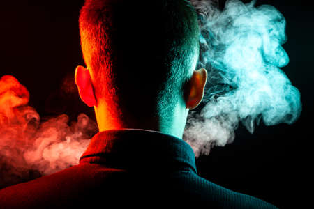 A view from the back on the head of a guy in a shirt smoking a vape and exhaling multi-colored smoke...