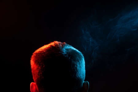 A view from the back on the head of a man in a shirt smoking a vape and exhaling multi-colored smoke of green at different directions from himself on a black isolated background. The thoughts and soul of human.の写真素材