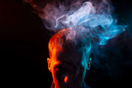 The face of a man in shadow and head is highlighted in red, who smokes a vape and gives out multi-colored smoke of blue and pink above his head, causing harm to his health. The thoughts and soul.の写真素材