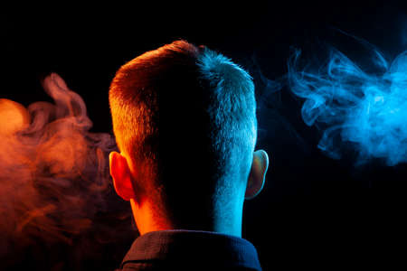 A view from the back on the head of a boy in a shirt smoking a vape and exhaling multi-colored smoke...