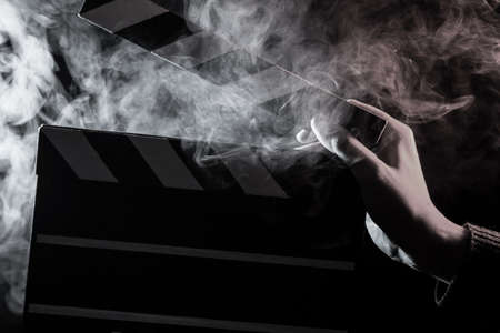 Close-up on an open clapperboard in hand before starting shooting a film with white smoke around with monochrome backlighting on a black isolated backgroundの写真素材