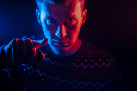 Portrait of a short-haired man with a shadow on a serious face dressed in a sweater with a colored backlight of blue and red points his finger at the viewer as Uncle Sam on a black isolated background.の写真素材