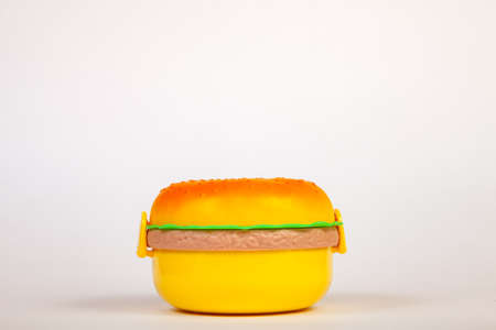 Lunchbox in the form of a yellow hamburger for preserving and carrying homemade food or sandwiches during working day. The symbol of artificial and unnatural harmful food on white isolated background.の写真素材