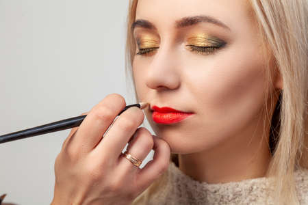 The make-up artist in the studio imposes a luxurious oriental make-up to a young girl blonde, in whose hand she brushes a lipstick of red color, with bright beige-gold and green shades in her eyes.のeditorial素材