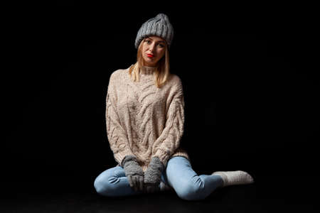 Young beautiful blonde woman in knitted gloves and hat in gray, blue jeans, beige sweater sits on the floor with crossed legs and looking away on a black isolated backgroundの写真素材