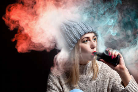 Blonde girl in a knitted sweater and hat holds an electronic cigarette vape in her hand with a manicure and exhales a lot of colored red and green smoke, with red lipstick on a black background.の写真素材