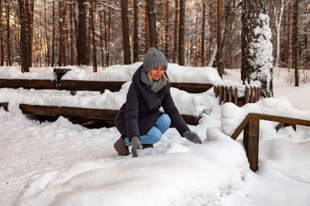 The blonde girl in a gray hat and gloves and a winter jacket squatted down and playing with the snow tossing it up in the forest on the background of a wooden bench.の写真素材