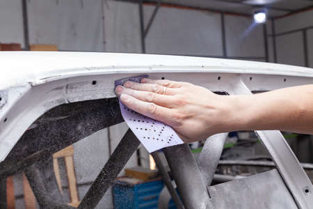 The body repairman grinds the white car's frame with purple emery paper in preparation for painting after applying putty in a vehicle repair workshop and auto service, dust crumbles down.の写真素材