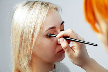 Visagiste with a brush in one hand putting makeup on the eyes of the model, the other hand pulls the...