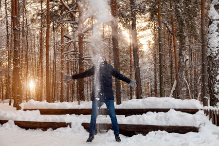 A young man in a winter coniferous forest is standing and catching with his hands the snow falling...