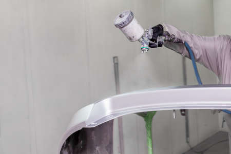 A male worker paints with a spray gun a part of the car body in silver after being damaged at an accident. Bumper from the vehicle during the repair in the workshop.の写真素材