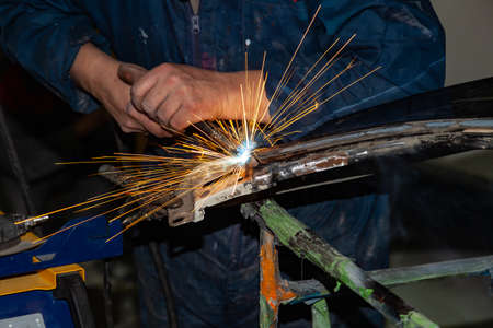 Close-up view strong man is a welder in blue working overalls without gloves on arms, a metal...