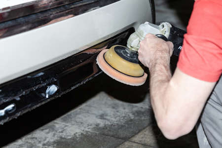 Close-up view on the hands of a male worker who holds a tool for polishing the bumper of a car while working in a vehicle detailing workshop. Repair part of the body by rotary polisher.の写真素材