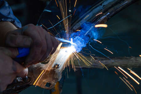 Close-up view strong man is a welder in working overalls without gloves on arms, a metal product is...