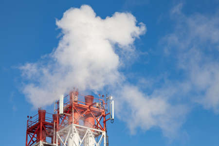 White harmful smoke coming out of the red-and-white pipes with mobile communication antennas at a factory in the city center against the background of a clear blue sky. Environmental problems.の写真素材