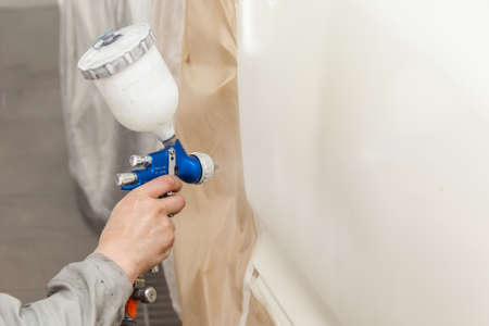 A male worker paints with a spray gun a part of the car body in white after being damaged at an accident. Door from the vehicle during the repair in the workshop. Auto service industry professionsの写真素材