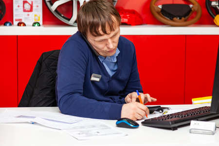 Novosibirsk, Russia - 04.25.19: Young male administrator talking on the phone at work, holding the phone to his ear and at the same time making notes on the paper with a pen sitting at a tableのeditorial素材