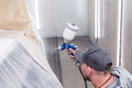 A guy worker paints with a spray gun a part of the car body in white after being damaged at an accident. Door from the vehicle during the repair in the workshop. Auto service industry professionsの写真素材