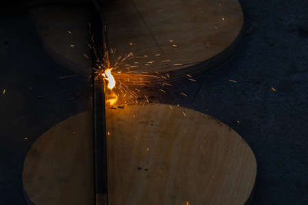 Close-up view strong man is a welder in blue working overalls without gloves on arms, a metal product is welded with a welding machine in the garage workshop, blue smoke and orange sparks fly awayの写真素材