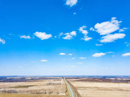 Aerial view of the asphalt road in a field with yellow grass during planting seeds and crops far from big cities on a clear spring day under a blue sky with trees to protect from the wind.の写真素材