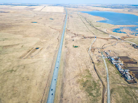 Aerial view of the asphalt road in a field with yellow grass during planting seeds and crops far from big cities on a clear spring day under a blue sky with trees to protect from the wind.の写真素材