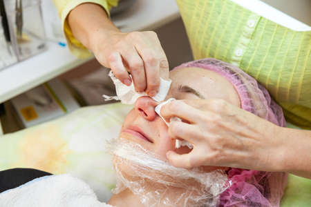 A young girl is lying on a couch during cosmetic procedures with a mask on the face above which beautician woman squeezes body fat and pimples with wipes on nose. Cleansing and make up.の写真素材