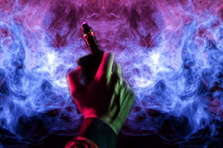 A man body part arm and hand view with green light in a orange hoodie smokes a vape and exhales a red smoke on bright blue and pink wavy background. Harm to human health and well-being.の写真素材