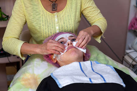 An experienced cosmetologist washes away a mask of white cream on the face of a young girl who lies on the couch during facial cleansing and beauty treatments. Cabinet cosmetologist, skin health.の写真素材