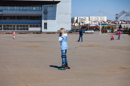 Leninsk Kuznetsky, Russia - 05.03.2019: A young blonde girl in a blue jacket is roller-skating around the square for walks and rest on a clear spring day.のeditorial素材