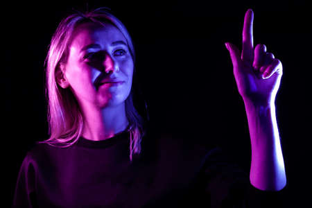 A young beautiful girl blonde highlighted in blue and pink on the sides raises her hand to the top and presses the power button on the touch screen and display. Modern technology and the futureの写真素材