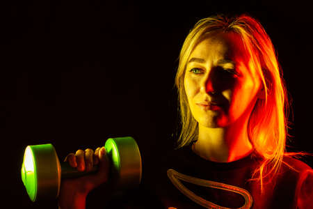 A young beautiful blonde girl in a black sweatshirt is holding a green plastic dumbbell in her hand...