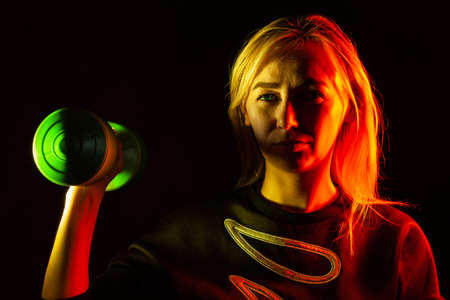 A young beautiful blonde woman in a black sweatshirt is holding a green plastic dumbbell in her hand raising up with backlight in red and yellow on the sides. Sports and healthy lifestyle.の写真素材