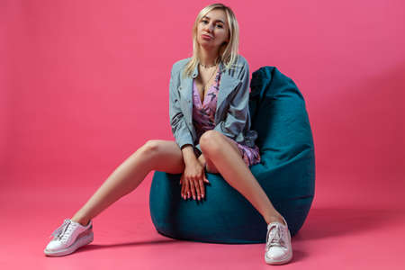 Beautiful blonde girl in a blue jacket sits on a green bag chair with her legs apart in the shape of...