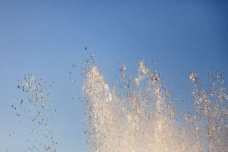 Close-up on splashes from the streams of an artificial fountain from which water flows in drops in the city park against the blue sky on a warm summer sunny dayの写真素材