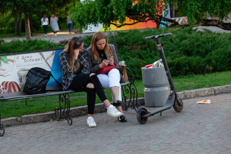 Novosibirsk, Russia - 05.28.2019: Two young girls are sitting on a bench and looking at a smartphone in a city park with green plants near a black electric scooterのeditorial素材
