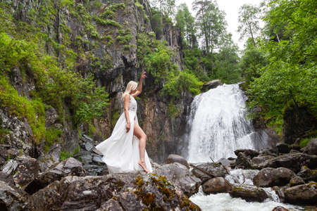 A young blond girl in an elegant pose bares her leg while reclining the hem of a boudoir dress, in the mountains against waterfall and stones, like a ballerina on a warm summer day in Altai green treeの写真素材