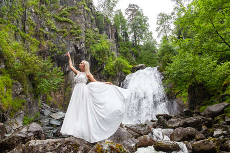 A young blonde girl in an elegant pose pulls up a boudoir dress in the mountains against a waterfall and stones raising her hands up like a ballerina on a warm summer day in Altai with green treesの写真素材