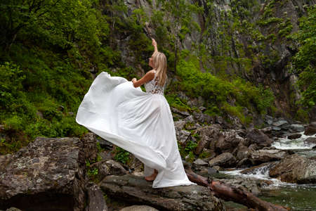 A young blonde girl in an elegant pose pulls up a boudoir dress up in the mountains against a waterfall and stones raising her hands up like a ballerina on a warm summer day in Altai with green treesの写真素材