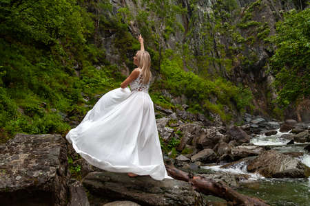 A young blonde girl in an elegant pose pulls up a boudoir dress up in the mountains against a waterfall and stones raising her hands up like a ballerina on a warm summer day in Altai with green treesの写真素材